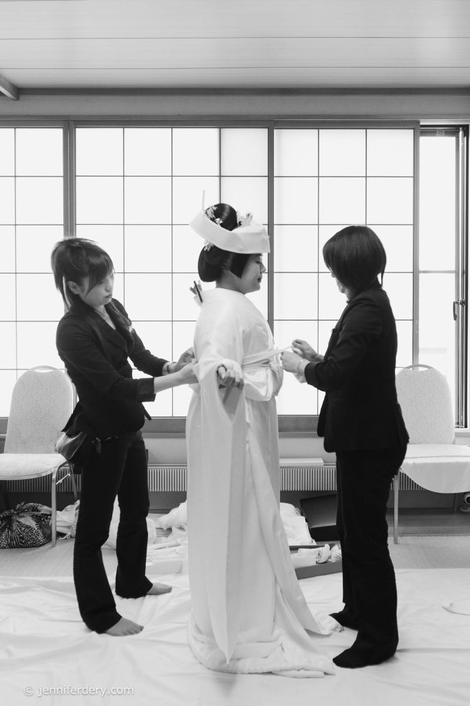 Two women help dress a bride in a traditional Japanese wedding kimono and headpiece in a bright room with shoji screens. The scene is calm and focused, with attention to detail in the attire.