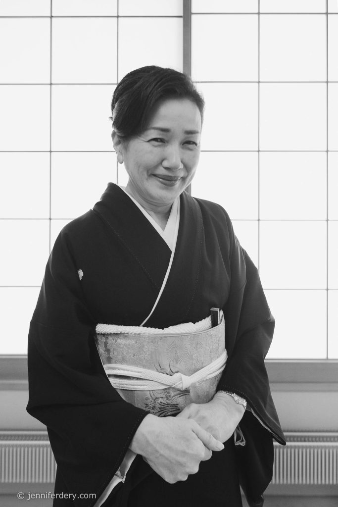 A woman dressed in a traditional kimono stands in front of a shoji screen, smiling gently with her hands folded. The black and white photo highlights the elegant patterns of her attire.