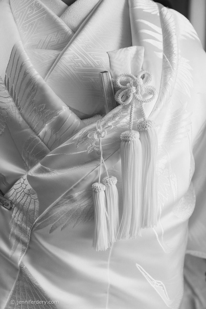 Close-up of a traditional Japanese wedding kimono in white silk with embroidered crane designs, featuring ornate knot and tassel details.