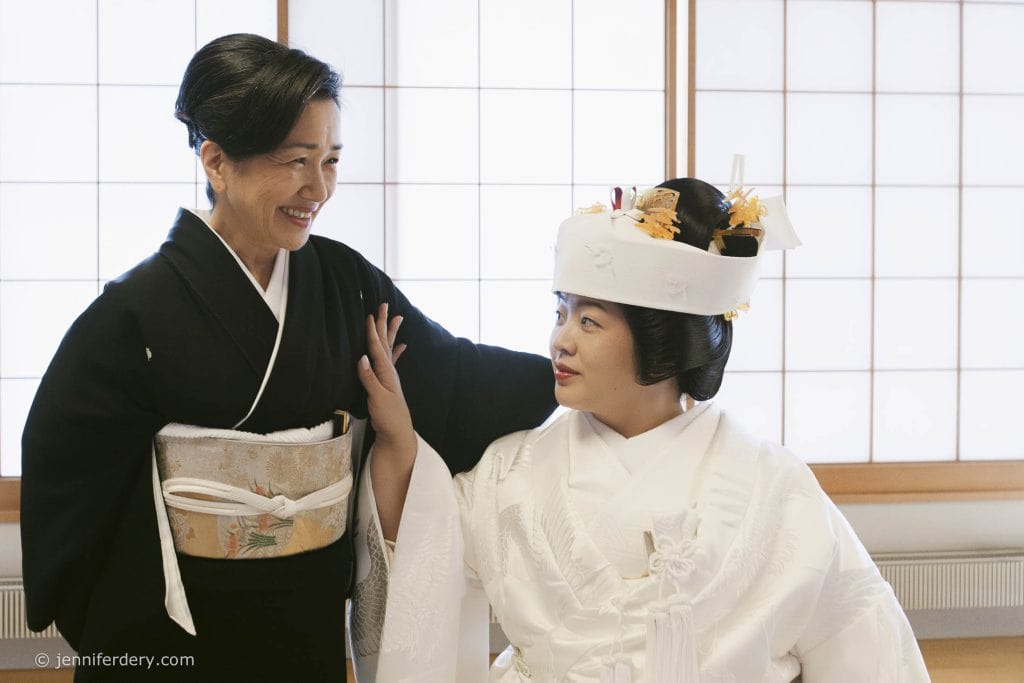 A woman in a black kimono smiles warmly at a bride in a traditional white Japanese wedding kimono and headpiece, inside a room with shoji screens and soft natural light.