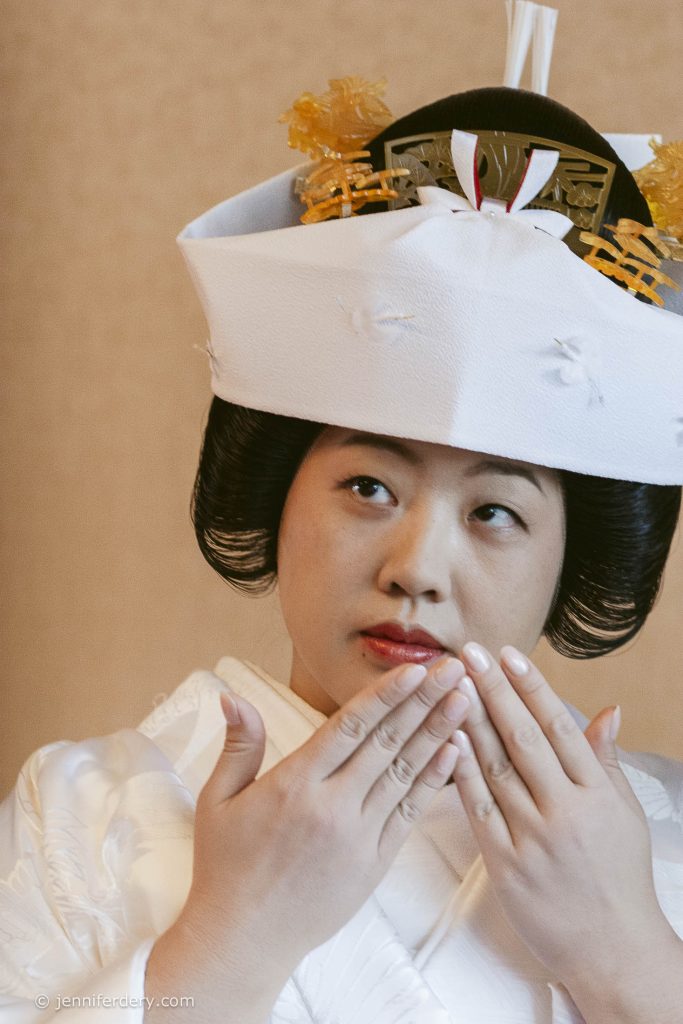 A woman dressed in a traditional Japanese bridal kimono and headdress gently touches her lips with her fingertips, looking slightly to the side.