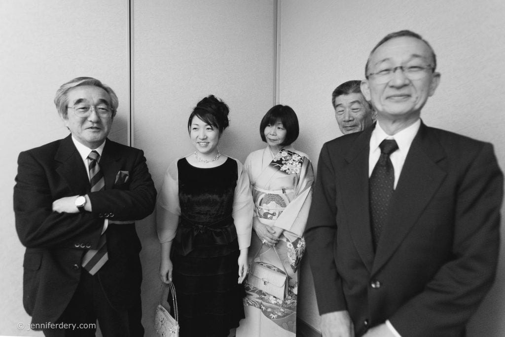 Five adults, three men in suits and two women in formal dresses, one in a kimono, stand together indoors, smiling and posing for the camera against a plain wall. The photo is in black and white.