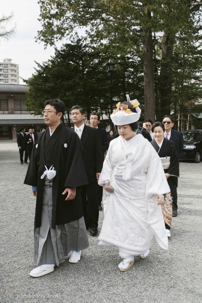 A bride and groom in traditional Japanese wedding attire walk outdoors, followed by family and friends. The bride wears a white kimono and headdress, and the groom wears a black kimono with striped hakama. Trees and buildings are in the background.
