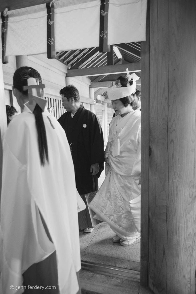 A Japanese bride in a traditional white wedding kimono and headpiece walks with a groom and attendants in ceremonial attire inside a wooden building.