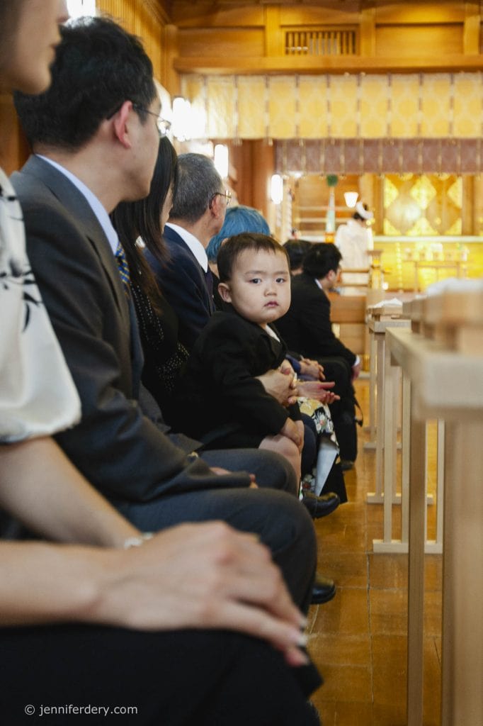 A young child in formal clothing sits on an adult’s lap among a row of people in a wooden hall, looking directly at the camera as a ceremony takes place at the front.