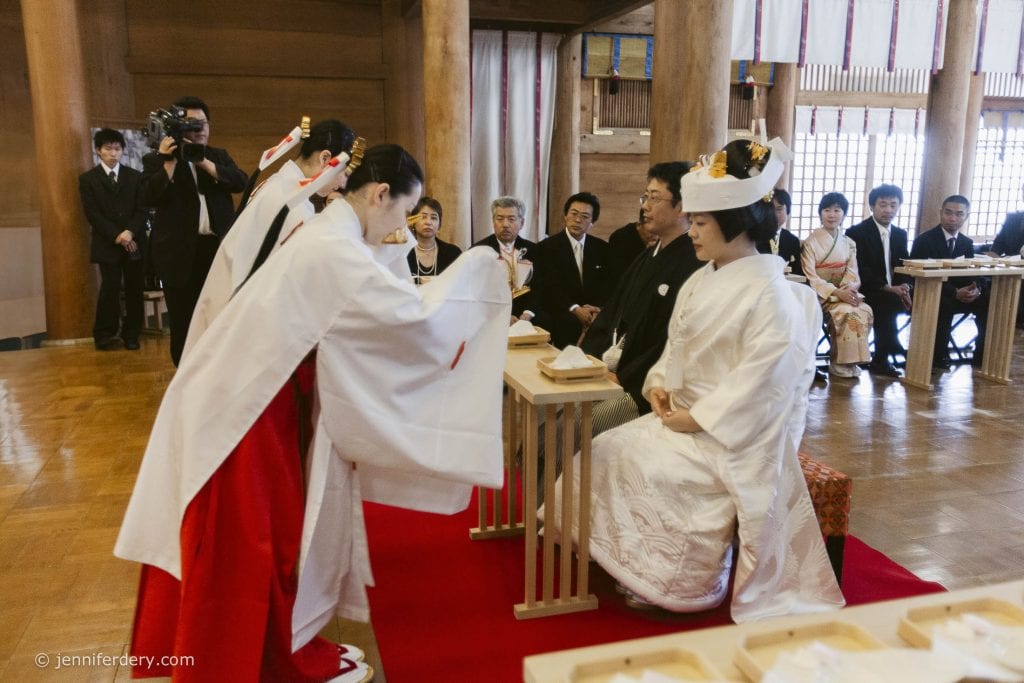 Two shrine maidens in white and red robes perform a ritual in front of a bride and groom dressed in traditional Japanese wedding attire, with guests seated in the background inside a wooden hall.