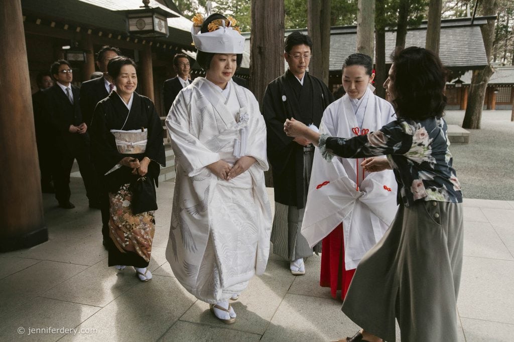 A bride in traditional white Japanese wedding attire walks with attendants and family members, all in formal dress, during a wedding ceremony at a shrine. A woman approaches them holding a small object.