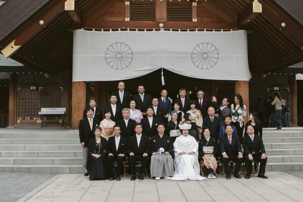 A large group of people in formal attire pose for a photo in front of a traditional Japanese shrine, with a bride in a white kimono and a groom in black seated at the center of the front row.