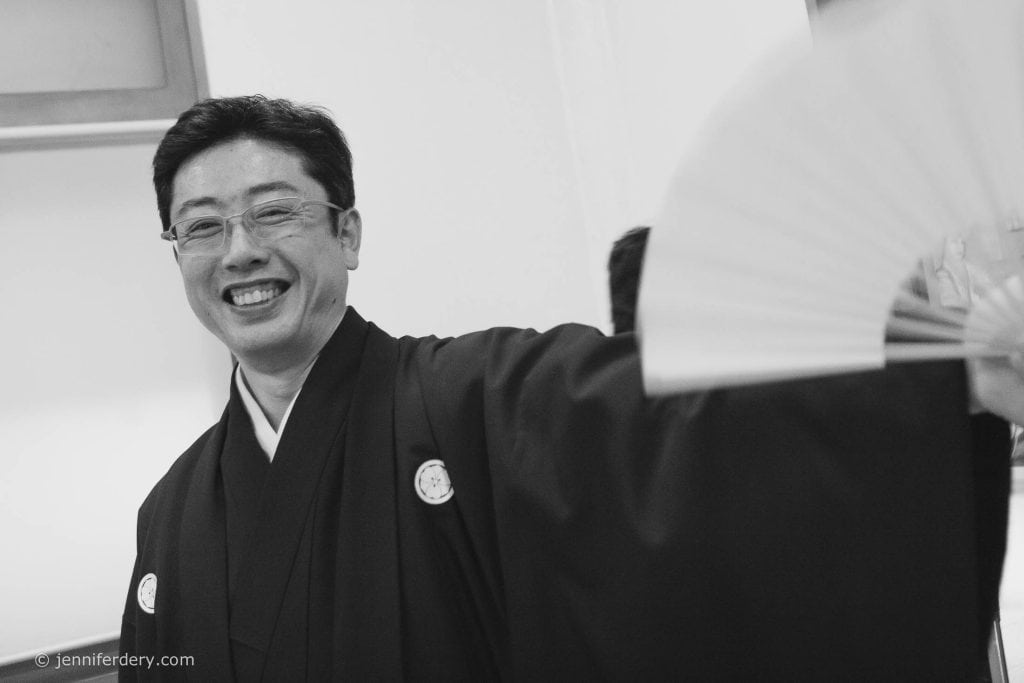 A man wearing traditional Japanese clothing smiles broadly while holding an open folding fan in front of him. The photo is in black and white.