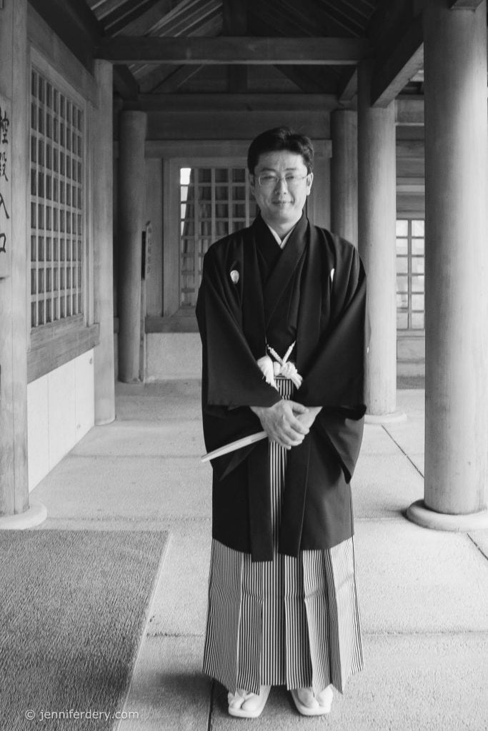 A man stands under a wooden temple walkway, wearing traditional Japanese formal attire including a kimono and hakama. He smiles slightly and holds a folded fan in his hands.