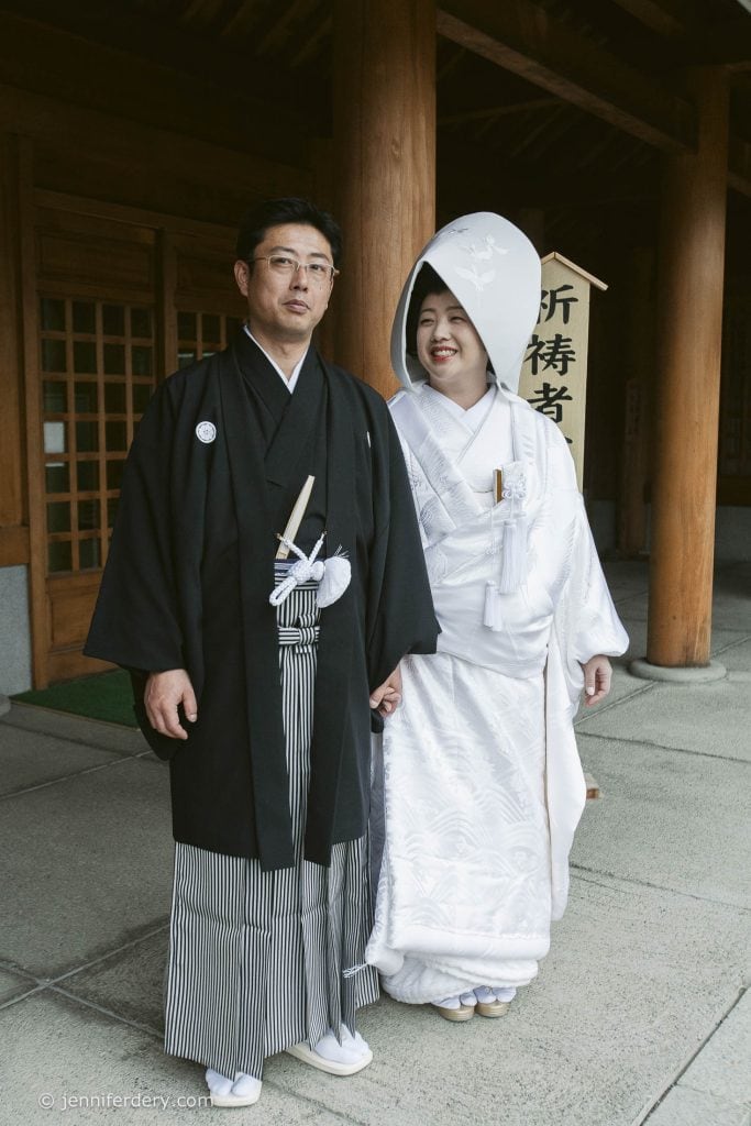 A couple in traditional Japanese wedding attire stands outside a wooden building. The groom wears a black kimono and striped hakama, while the bride wears a white kimono and a tall, rounded headpiece, smiling as they hold hands.