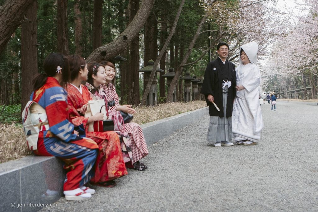 A bride and groom in traditional Japanese wedding attire stand together under cherry blossoms, while six women in colorful kimonos sit nearby, smiling and watching them. Tall trees line the path in the background.