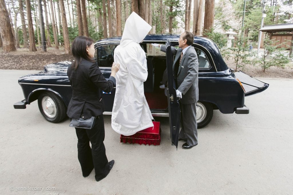 A bride in a traditional white Japanese wedding kimono steps into a black car, assisted by two people dressed in black suits, surrounded by tall trees in an outdoor setting.