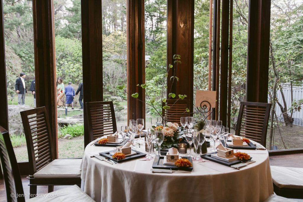 A round dining table set for a formal event with elegant tableware, glasses, and floral centerpieces, positioned by large windows overlooking a garden where several people are gathered.