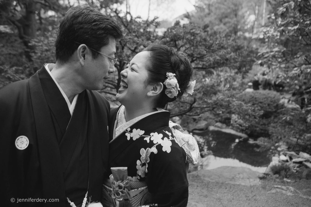 A couple in traditional Japanese attire smile and laugh together outdoors, surrounded by lush greenery and a small stream in the background.