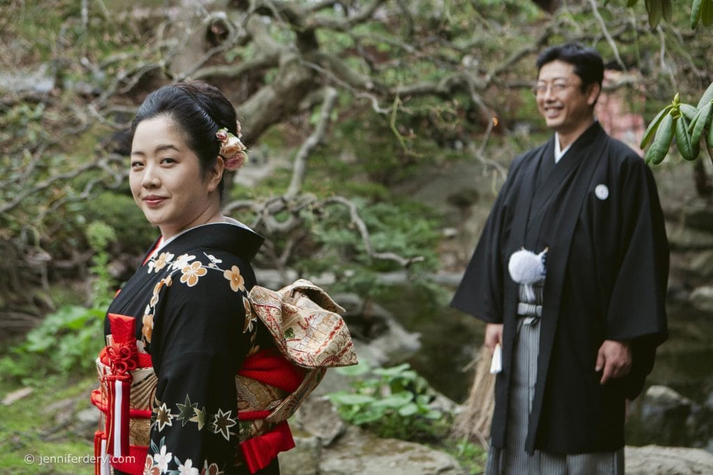 A woman in a black kimono with floral patterns stands in the foreground, smiling. Behind her, a man in traditional Japanese attire stands near greenery and a tree. Both appear happy and outdoors.