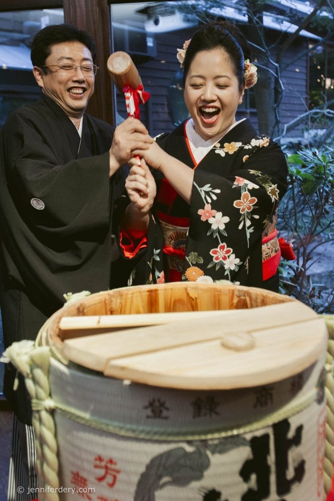 A smiling couple in traditional Japanese attire joyfully break open a sake barrel with a wooden mallet during a celebration. The woman wears a floral kimono and the man supports her hand as they share the moment.
