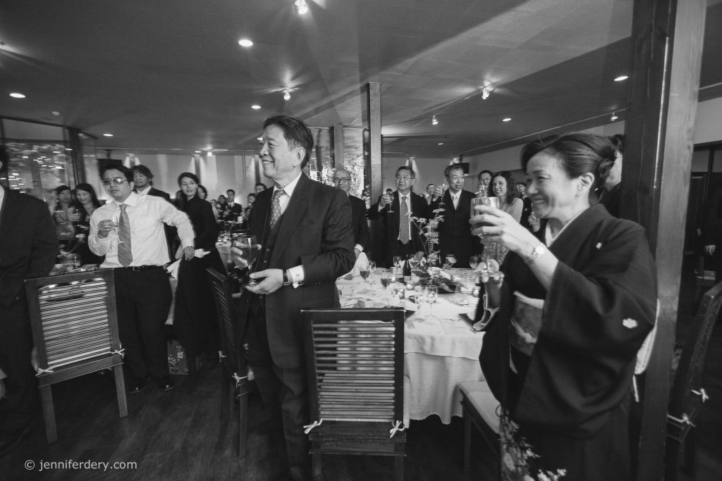 A group of well-dressed people, including a man in a suit and a woman in a kimono, stand and raise glasses at a formal event or celebration in a decorated dining room.
