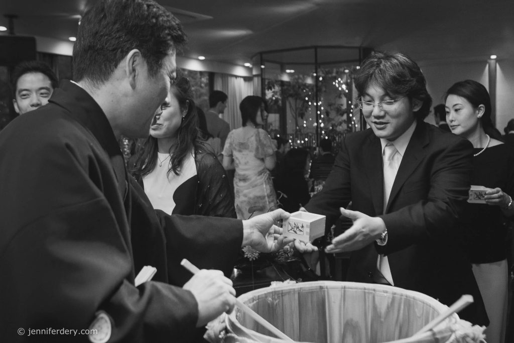 A man in a suit receives a small wooden box from another man in traditional Japanese attire during a lively indoor celebration. People in the background are smiling and observing the exchange.