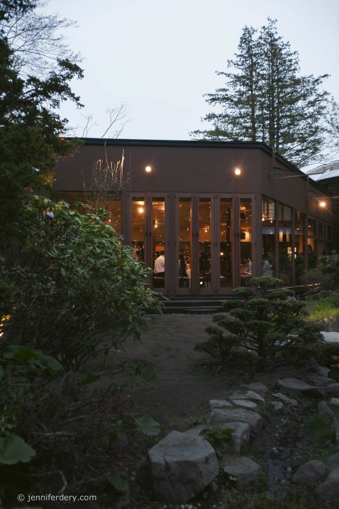 A modern building with tall windows glows warmly at dusk, surrounded by lush greenery and a stone path. Trees and shrubs frame the scene, creating a peaceful outdoor atmosphere.