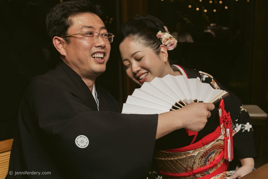 A smiling couple in traditional Japanese attire laugh together. The man holds a white folding fan, while the woman, wearing a floral hair accessory, leans in close. Both appear joyful and engaged in a celebratory moment.