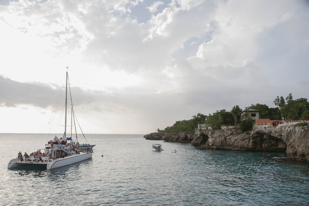 Rockhouse Hotel, Negril, Jamaica: A large catamaran with people aboard sails near a rocky coastline with houses and greenery, under a cloudy sky at sunset. A small boat and swimmers are nearby in the calm blue water.