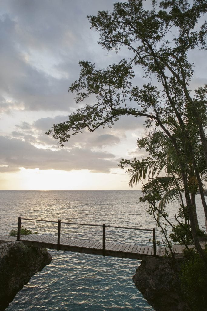 Rockhouse Hotel, Negril, Jamaica: A wooden footbridge crosses over turquoise water between rocks, with trees and palm fronds framing a view of the ocean at sunset under a partly cloudy sky.
