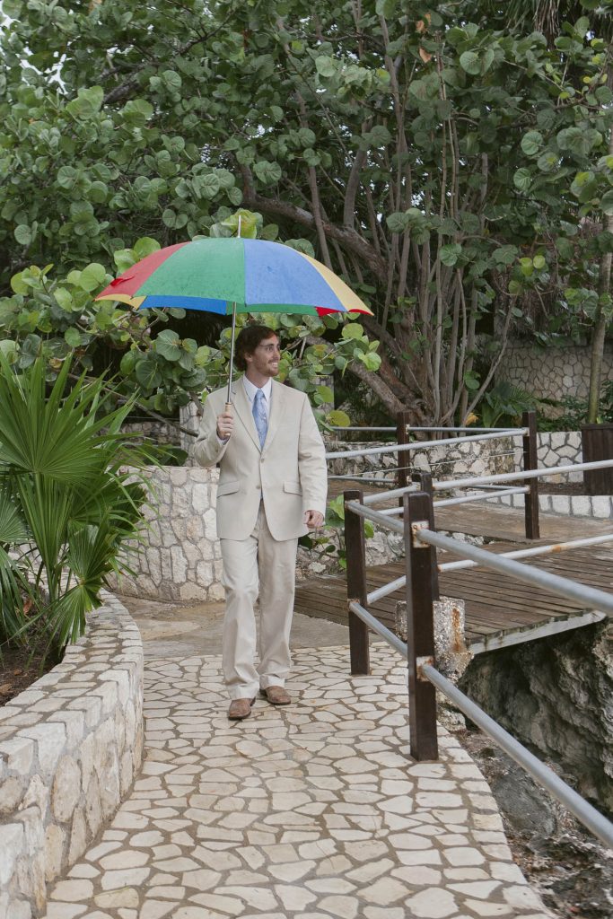 Rockhouse Hotel, Negril, Jamaica: A man in a beige suit and blue tie walks on a stone path holding a colorful rainbow umbrella, surrounded by lush green trees.