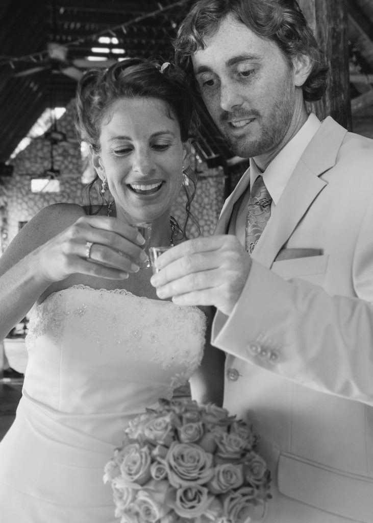 A bride and groom in wedding attire smile as they raise shot glasses together, holding a bouquet of roses. The photo is in black and white and appears to be taken at a rustic indoor venue.