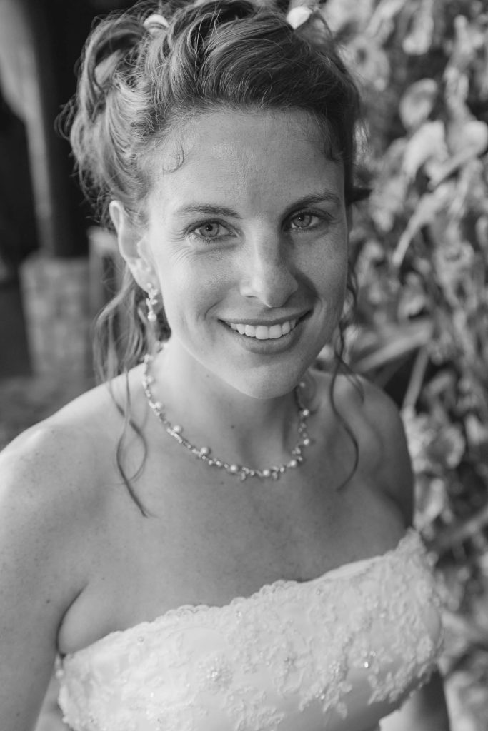 A woman in a strapless, embroidered wedding dress smiles at the camera. She has wavy hair styled up, wears a necklace and earrings, and stands in front of leafy greenery. The photo is in black and white.
