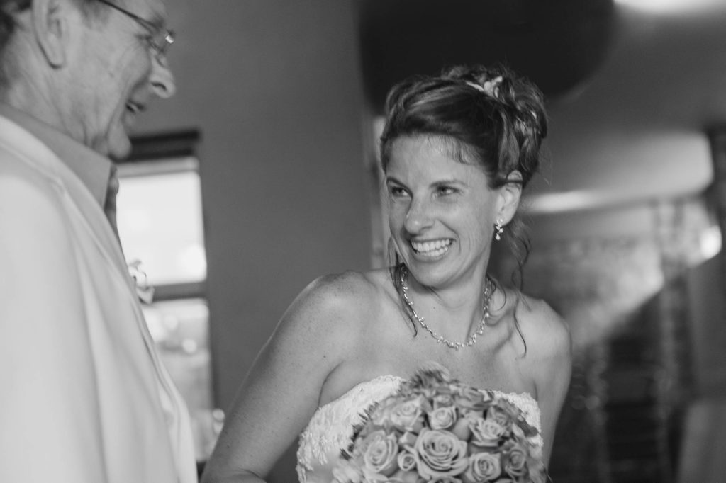 A bride in a strapless wedding dress smiles joyfully while holding a bouquet of roses, looking at an older man in glasses who is also smiling. The image is in black and white.