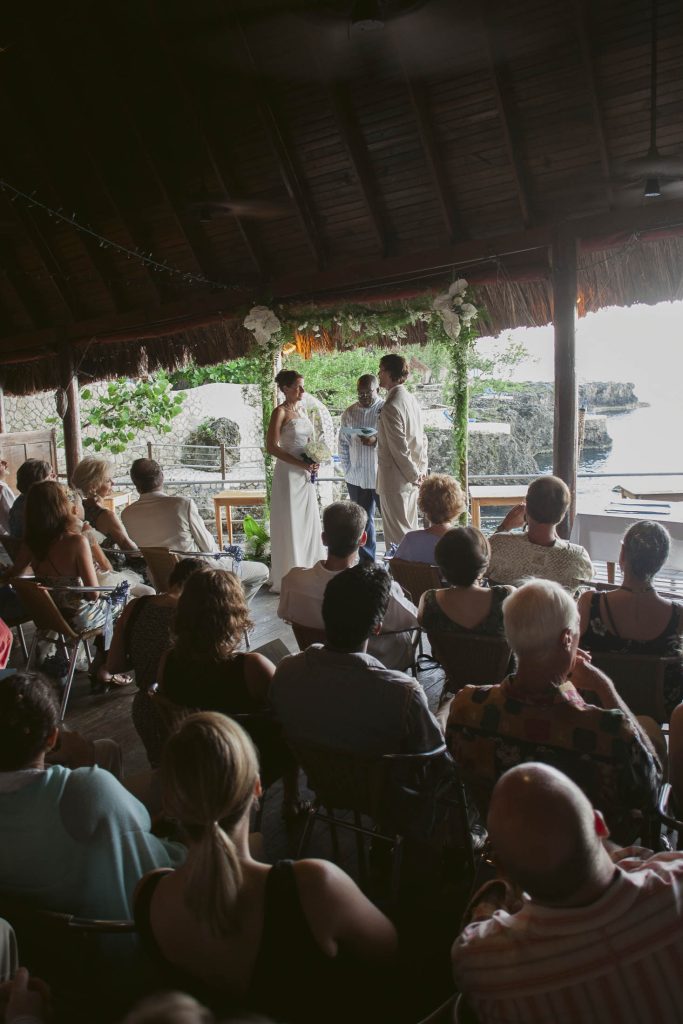 Rockhouse Hotel, Negril, Jamaica: A bride and groom stand together under a rustic pavilion, exchanging vows before guests seated inside, with greenery and water visible outside through open walls.