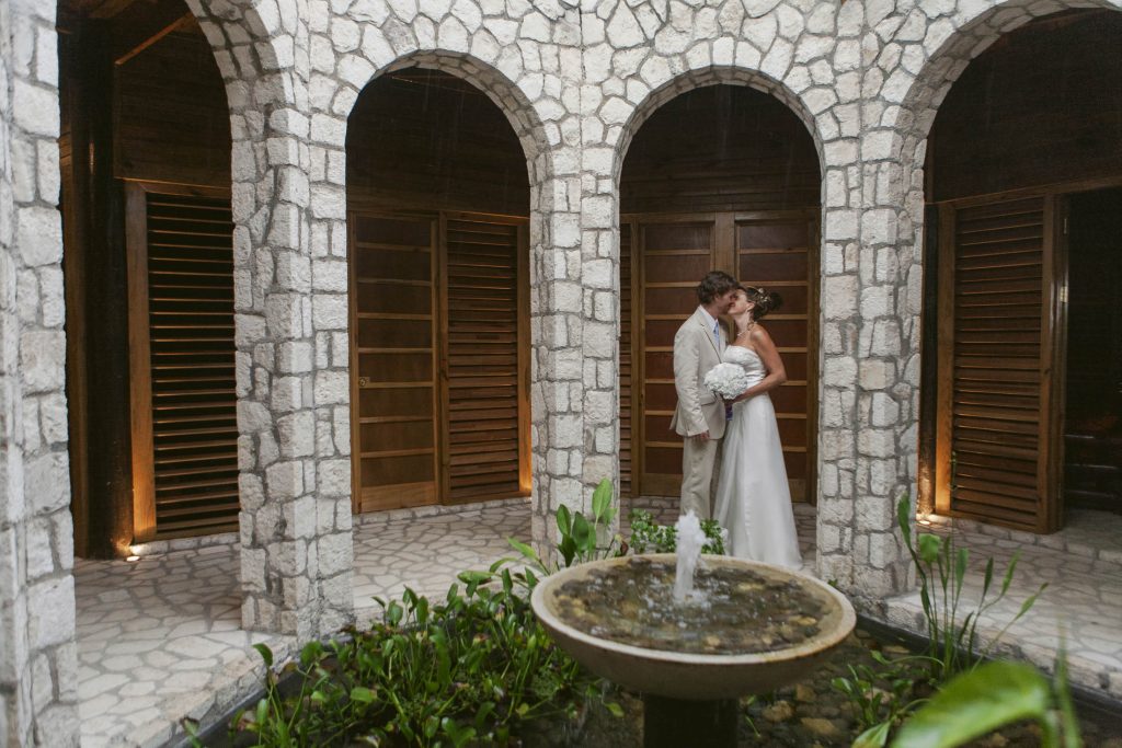 Rockhouse Hotel, Negril, Jamaica: A bride and groom embrace in front of a stone archway, surrounded by wooden shutters and a small fountain with green plants in the center. The setting has a romantic, rustic atmosphere.