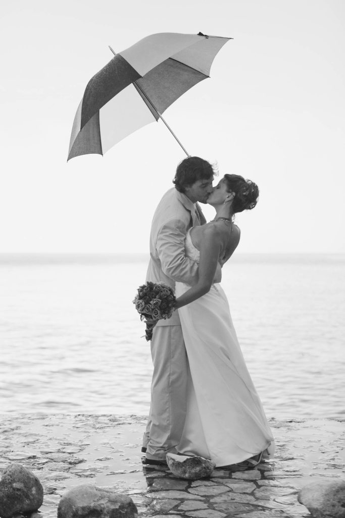 Rockhouse Hotel, Negril, Jamaica: A bride and groom stand on a stone path by the water, sharing a kiss under an umbrella. The bride holds a bouquet of flowers, and both are dressed in formal wedding attire. The image is in black and white.
