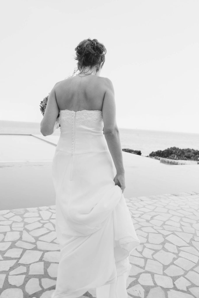 Rockhouse Hotel, Negril, Jamaica: A bride in a strapless white gown walks away on a stone path, holding her dress with one hand. The scene overlooks water and rocky terrain in the background. The image is in black and white.