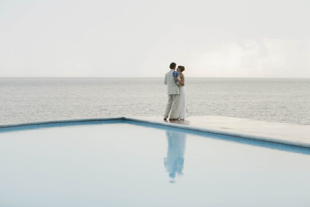 Rockhouse Hotel, Negril, Jamaica: A couple dressed in wedding attire stands close together at the edge of a pool, facing the ocean under a pale sky. Their reflection is visible in the calm pool water in the foreground.
