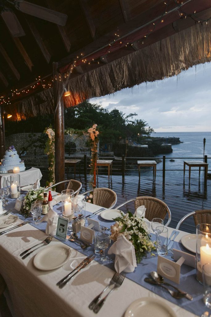 Rockhouse Hotel, Negril, Jamaica: A beautifully decorated outdoor dining table set for a formal event overlooks the ocean at sunset. Candles, flowers, and a wedding cake are on the table under a thatched roof with string lights.