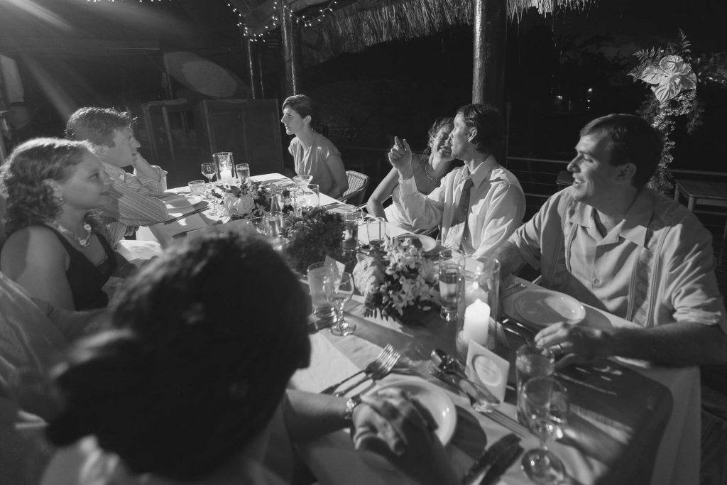 A group of people dressed in semi-formal attire sit around a decorated outdoor dinner table at night, talking and enjoying each other’s company under string lights.