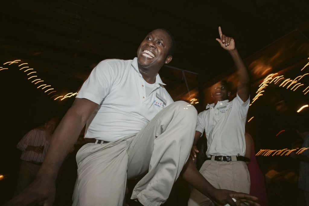 Two men in light-colored shirts and pants dance energetically at night, one smiling broadly with his knee raised and the other pointing upward, with string lights blurred in the background.