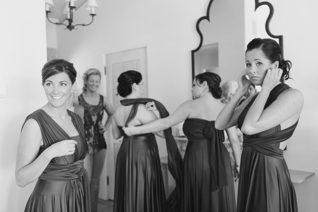 Four women in long dresses gather in a room, two adjusting their outfits, one smiling, and another helping fasten a dress. A large mirror and chandelier are visible in the background. The scene appears joyful and busy.