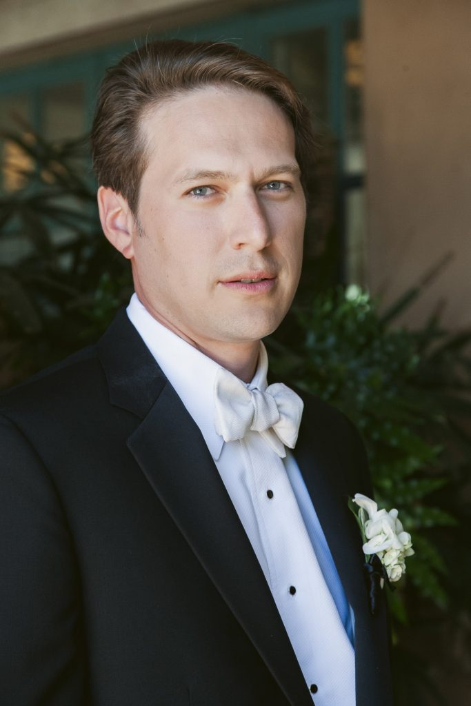 A man in a black tuxedo with a white shirt and bow tie stands indoors, looking at the camera with a neutral expression. He has short brown hair and a boutonnière pinned to his jacket. Green plants are in the background.