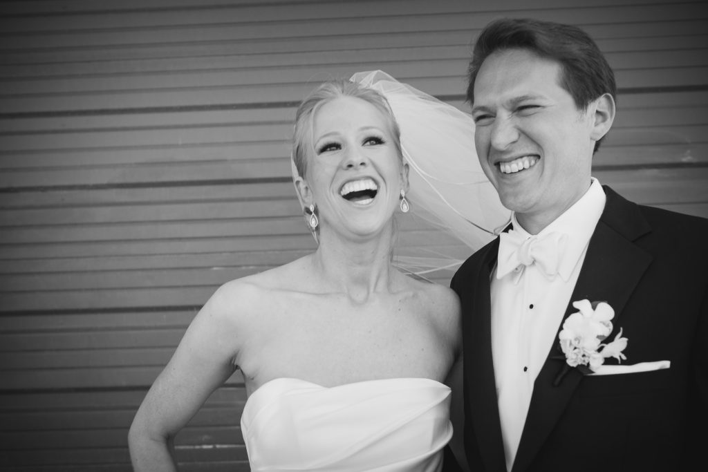 A bride and groom, both smiling and laughing, stand together in front of a striped wall. The bride wears a strapless dress and veil; the groom is in a tuxedo with a boutonniere. The mood is joyful and celebratory.