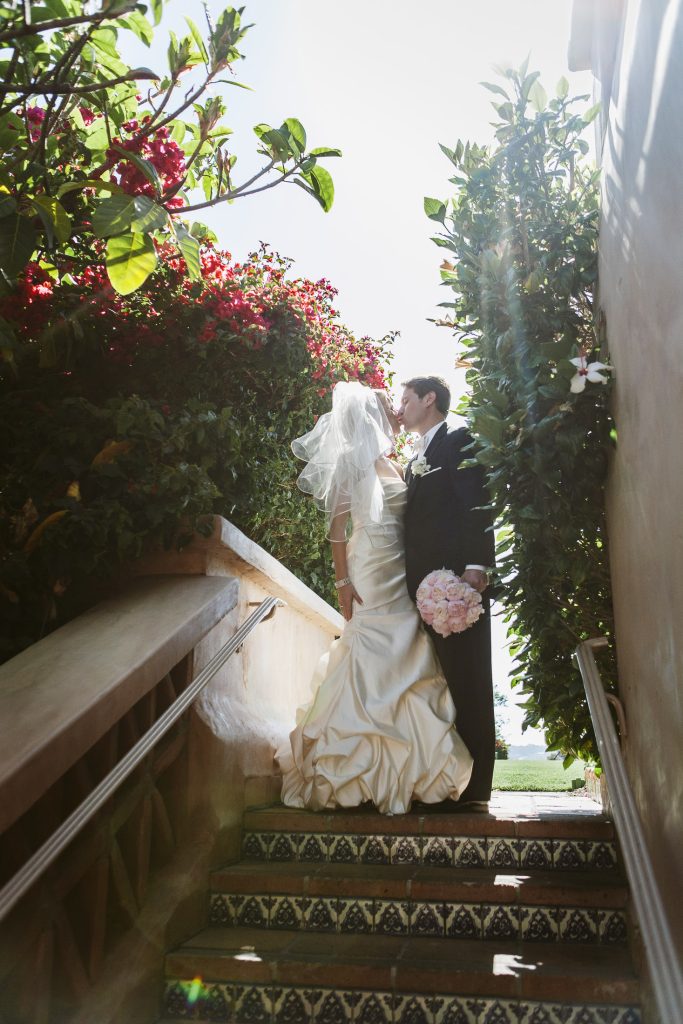 A bride and groom stand on tiled stairs surrounded by lush greenery and flowers, sharing a kiss in sunlight. The bride holds a bouquet and wears a veil; the groom is in a black suit.