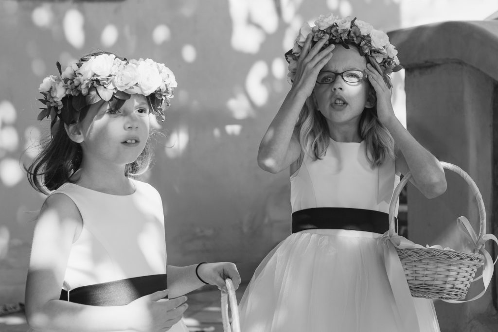 Two young girls in white dresses and flower crowns stand outside in sunlight, holding baskets. One girl looks forward calmly, while the other adjusts her glasses with both hands, appearing surprised or flustered.