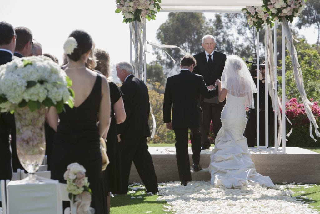 A bride in a white gown and veil walks up an aisle covered with white petals, accompanied by a man in a suit, toward an officiant waiting under a floral arch at an outdoor wedding ceremony, while guests look on.