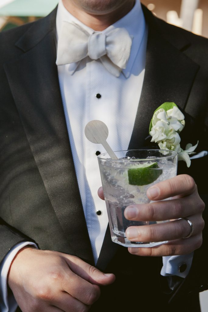 A man in a tuxedo with a white bow tie holds a clear cocktail garnished with lime and a stir stick. He wears a boutonnière with white flowers and a wedding band on his left hand.