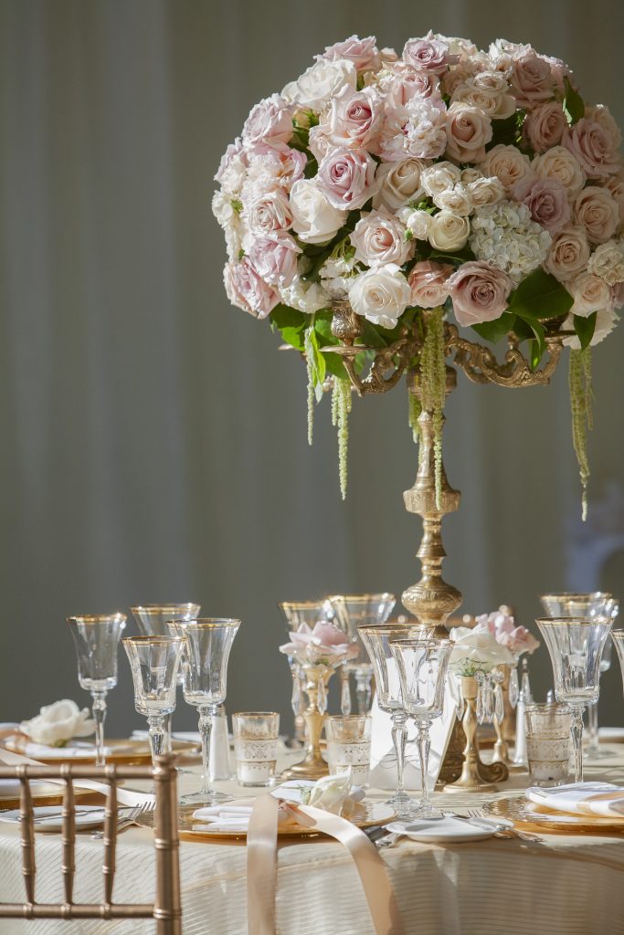A formal table setting with gold-rimmed glassware, gold plates, and an ornate centerpiece featuring a large bouquet of pink and white roses on a pedestal. Soft lighting creates an elegant atmosphere.