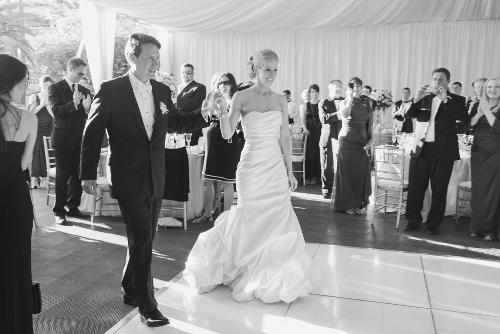 A bride in a strapless white gown and a groom in a tuxedo walk hand in hand into a wedding reception, smiling as guests stand and applaud around them. The setting is bright and elegant with draped white fabric.