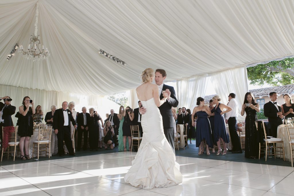 A bride and groom share their first dance under a white draped tent, surrounded by elegantly dressed guests watching and taking photos.