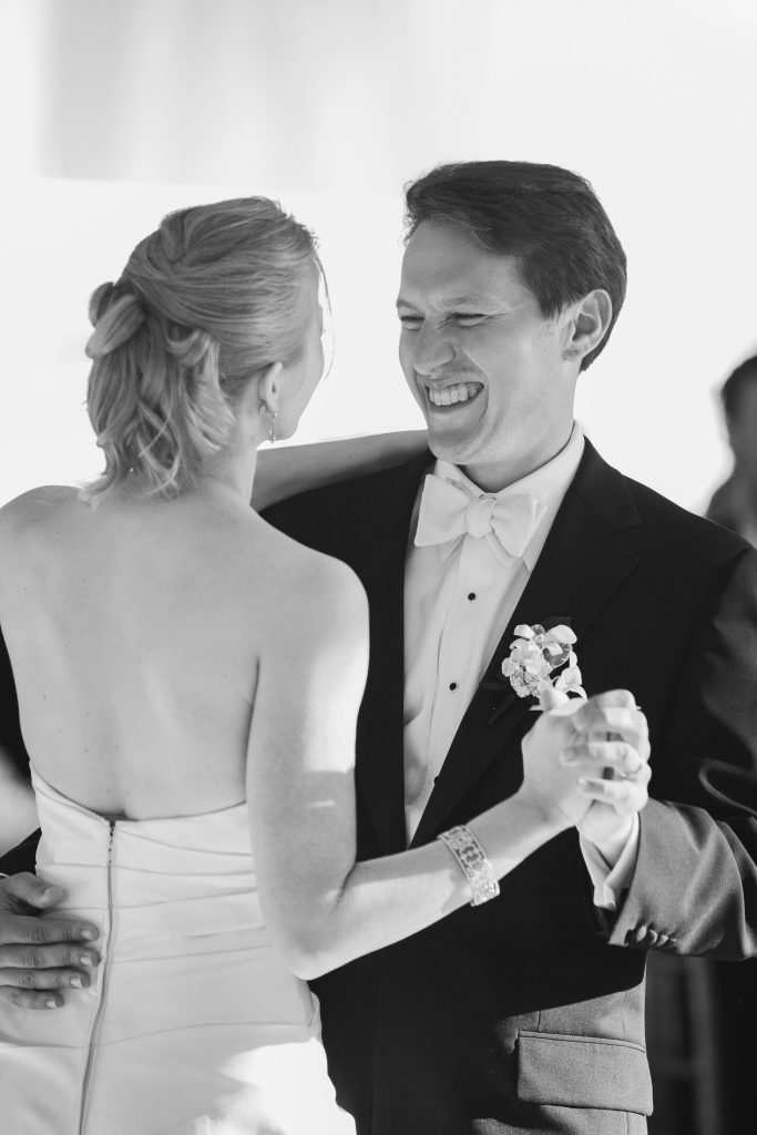 A bride and groom smile at each other while dancing together, dressed in formal wedding attire. The groom wears a tuxedo with a bow tie and boutonniere, while the bride wears a strapless gown.